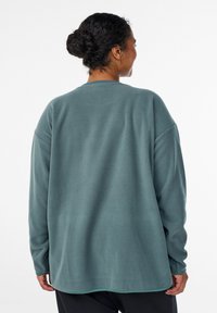 Fleecový pulóver v tyrkysovej farbe, s voľným strihom, dlhými rukávmi a zaobleným lemom. Mäkká textúra s minimálnymi švami a bez viditeľných zapínaní.