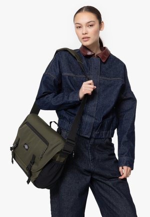 Green and black messenger bag with front pocket, adjustable strap, and buckle closures. Model wearing denim jacket and trousers.