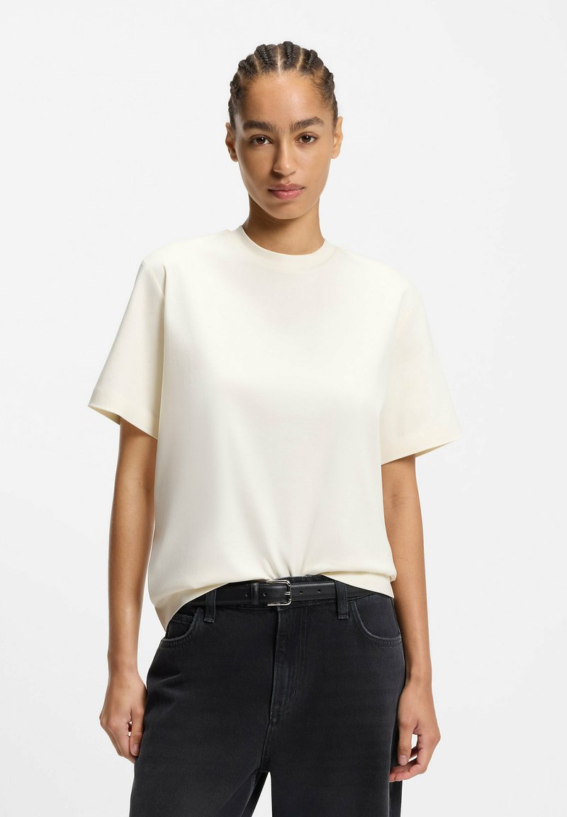 Haut crème à manches courtes en tissu lisse, doté d'un col rond et d'une coupe décontractée, associé à un jean en denim foncé et une ceinture noire.