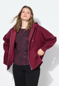 Sudadera con cremallera burdeos con bolsillos, sobre una camiseta negra que presenta un patrón de rayas de cebra rosa. La tela parece suave y texturizada.