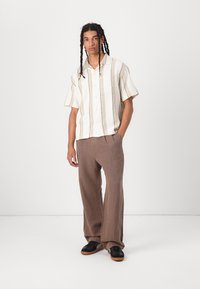 Chemise à manches courtes en crème avec des rayures verticales marron, associée à un pantalon large marron et des baskets noires. Design simple, coupe décontractée.