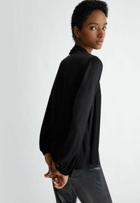Blusa nera a maniche lunghe con design plissettato, tessuto leggero e polsini arricciati. Sul retro presenta un collo alto e una texture liscia.