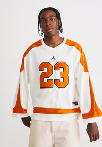Man with braided hair wearing white and orange sports jersey with number 23, standing against plain white background.