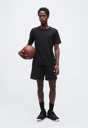 Νεαρός άνδρας με μαύρη αθλητική φόρμα κρατώντας ένα μπάσκετ στο ύψος της μέσης, στέκεται μπροστά σε απλό φωτεινό φόντο.