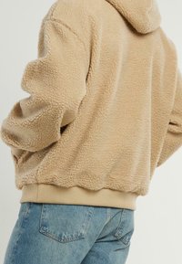 Sudadera de forro polar beige con un material texturizado y suave, que cuenta con capucha, puños y dobladillo acanalados. Combinada con jeans de mezclilla azul claro.