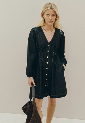 Donna con capelli biondi indossa un abito nero abbottonato con maniche lunghe, tenendo una grande borsa marrone scuro su uno sfondo semplice.