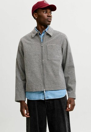 Homme portant une casquette rouge, une veste zippée grise à carreaux, une chemise en denim bleue en dessous et un pantalon noir regardant vers sa gauche.