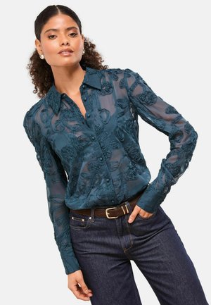 Türkisfarbige, durchsichtige Bluse mit floralen Stickereien, Knopfverschluss, langen Ärmeln und spitzen Kragen; kombiniert mit dunklen Jeans und einem braunen Gürtel.