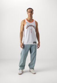 Camiseta de baloncesto blanca con acentos naranjas, con el texto "SAN ANTONIO", número 1, combinada con unos vaqueros holgados de color azul claro y zapatillas blancas.