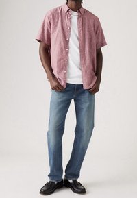 Camisa de boton corto de manga, color rojo claro, sobre una camiseta blanca; combinada con jeans azules y zapatos negros. Patrones simples y corte clásico.