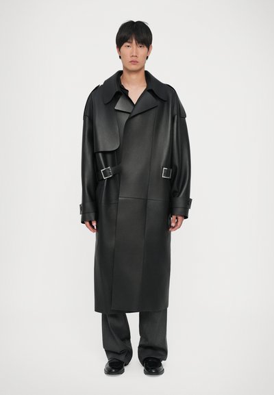 Manteau trench en simili cuir noir avec un grand col, des détails en boutons et des accents de boucle ; associé à un pantalon large gris et des chaussures noires.