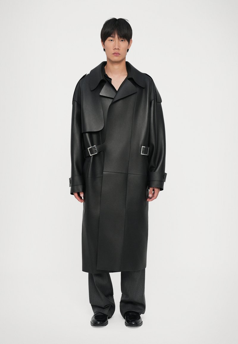 Sort trenchcoat i kunstlæder med oversized krave, knapdetaljer og spænde aksenter; parret med brede grå bukser og sorte sko.