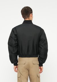Giubbotto bomber nero con colletto alto e polsini a costine. Presenta un tessuto liscio e lucido, con una vita aderente e una silhouette rilassata.
