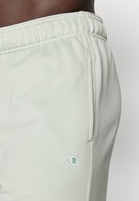Light green cotton sweatpants with an elastic waistband, side pockets, and a subtle embroidered logo on the left thigh. Smooth texture.