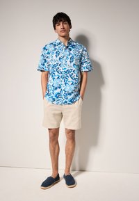 Chemise à manches courtes bleue à motif floral avec des accents blancs, shorts beige et chaussures slip-on bleu marine. Tissu léger avec une coupe décontractée.