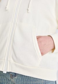Krem fôret med glidelås med snorer, som har en frontlomme og ribbede mansjetter, parret med denimjeans. Myk, glatt stofftekstur.