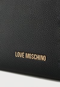 Μαύρη υφασμάτινη δερμάτινη επιφάνεια με χρυστά μεταλλικά γράμματα που σχηματίζουν τη φράση «LOVE MOSCHINO» στο κέντρο κοντά στο κάτω μέρος.