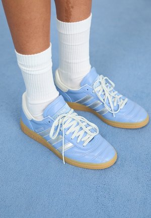 Lyseblå lær-sneakers med hvite detaljer, flate snører og gummisåle, kombinert med hvite ribbestrikkede sokker på en blå overflate.