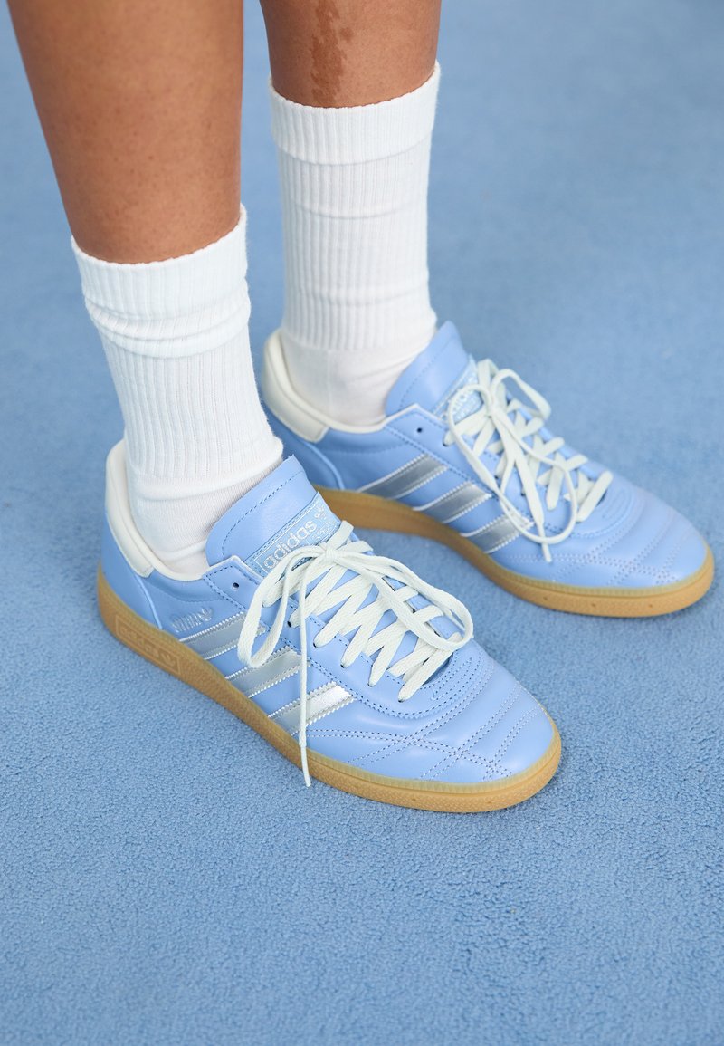 Svetlomodré kožené tenisky s bielymi detailmi, plochými šnúrkami a gumenou podrážkou, spárované s bielymi rebrovanými ponožkami na modrom povrchu.