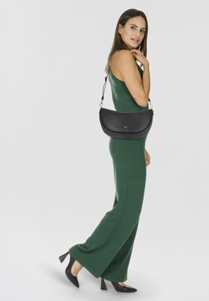Bolso de hombro de cuero negro con forma curved, con una superficie texturizada y herrajes en tono dorado, usado con un mono verde.
