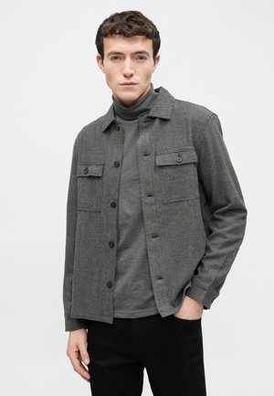 Gray checkered jacket with two chest pockets and black buttons, worn over a dark turtleneck; features long sleeves and a relaxed fit.