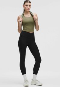 Donna con un top senza maniche verde oliva e leggings neri, che tiene un asciugamano verde attorno al collo, indossando scarpe da ginnastica e calze bianche.