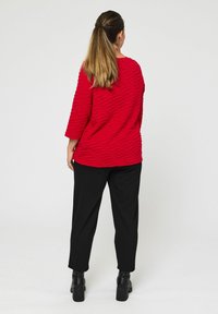 Rode gestructureerde top met golfpatronen, driekwartmouwen; gecombineerd met zwarte broeken en stevige zwarte laarzen. Witte achtergrond.