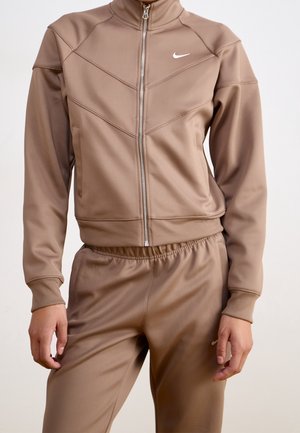 Personne portant une veste zippée Nike marron et un pantalon assorti à taille élastique, debout sur un fond neutre.