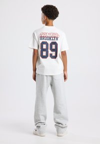 Valkoinen puuvillapaita, jossa on suuri "BROOKLYN" -teksti ja numero "89" tummansinisellä, yhdistettynä harmaisiin verkkareihin. Malli näkyy takaa.
