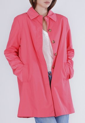 Kobieta ubrana w jaskraworóżowy płaszcz na guziki z rękami w kieszeniach, białą bluzkę w wzory i niebieskie dżinsy na jednolitym tle.