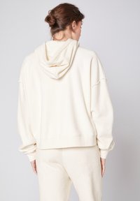 Haut à capuche en molleton crème, doux et confortable, avec une coupe décontractée, des épaules tombantes et une grande capuche. Texture douce et design cropped au niveau de la taille.