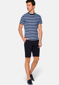 Pasiasta koszulka w kolorze granatowym i jasnoniebieskim, okrągły dekolt. Połączona z ciemnymi szortami i granatowymi sneakersami. Modelka stoi, ręce swobodnie.