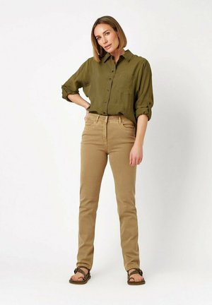 Frau steht mit der Hand auf der Hüfte, trägt ein olivgrünes Hemd mit Knöpfen, beige Hose und braune Sandalen vor einem schlichten weißen Hintergrund.