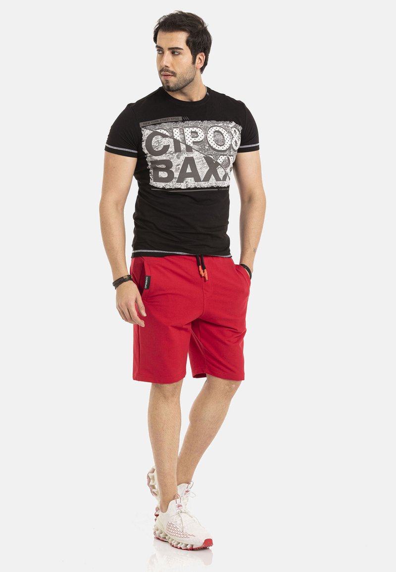 Zwarte T-shirt met korte mouwen en witte grafische tekst, gecombineerd met rode shorts met zijzakken en een verstelbaar trekkoord. Witte sneakers.