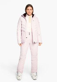 Helle rosa isolierte Skijacke mit Reißverschluss, kombiniert mit passenden Hosen, die Knöpfe an der Taille und ein Logo-Detail in der Nähe des Knöchels aufweisen.