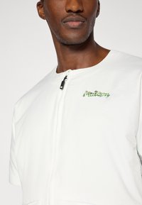 Λευκό μπουφάν με φερμουά ρ και κοντά μανίκια, με πράσινα κεντημένα γράμματα στο στήθος και υφή ελαφριάς, υφασμάτινης επιφάνειας.