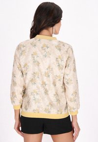 Blommönstrad bomberjacka i ljusbeige med gula ribbade manschetter och nederkant. Texturerad yta med spridda gula och gröna blommönster.