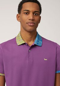 Lila polo skjorta med mönstrad krage i blått och grönt; har en liten gul hundlogotyp på bröstet. Bomullsmaterial, korta ärmar.