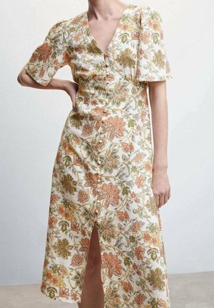 Femme portant une robe midi blanche avec un imprimé floral orange, vert et jaune, des manches courtes bouffantes, des boutons sur le devant et une fente frontale.