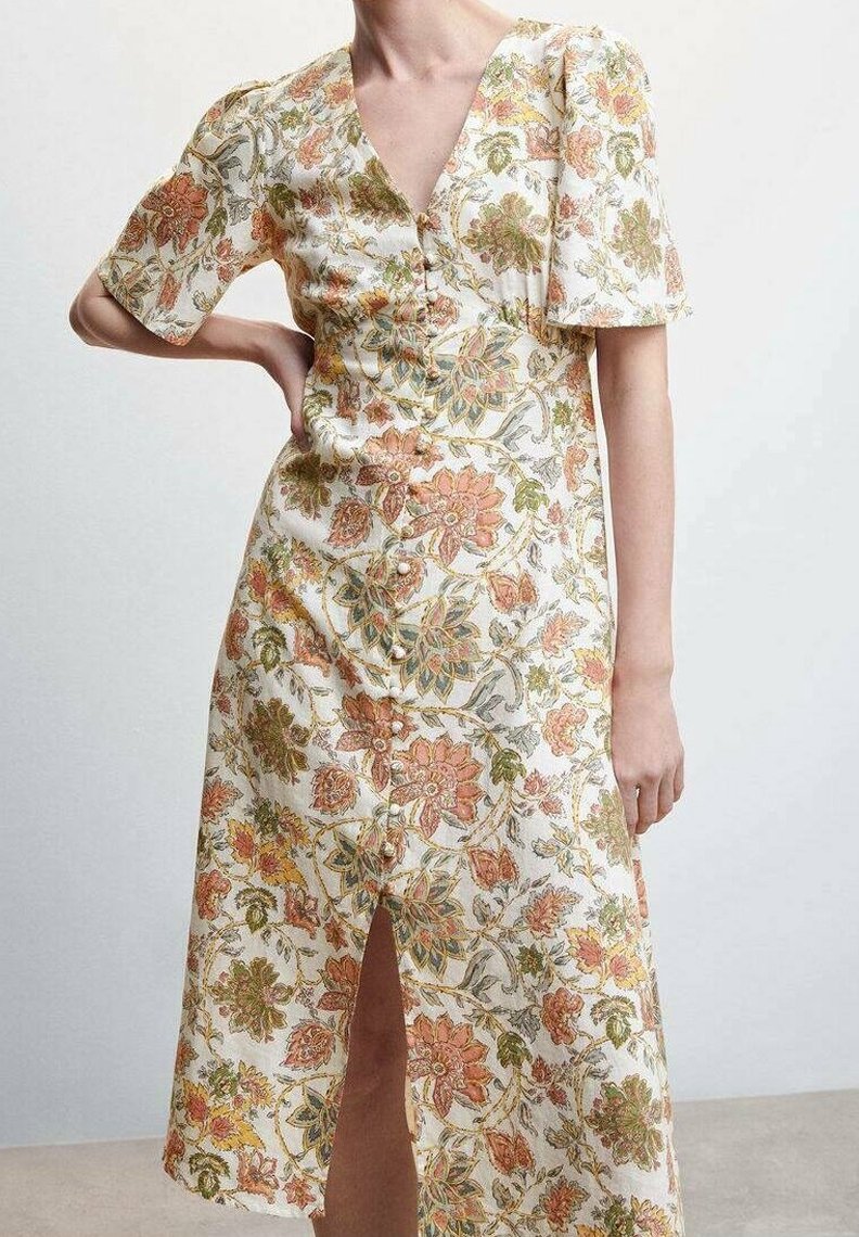 Femme portant une robe midi blanche avec un imprimé floral orange, vert et jaune, des manches courtes bouffantes, des boutons sur le devant et une fente frontale.