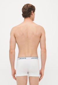 Uomo visto di spalle che indossa boxer bianchi Lacoste con cintura logata nera su sfondo semplice.