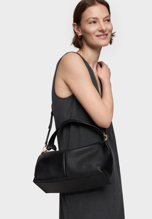 Mujer con vestido oscuro sin mangas que lleva un bolso de cuero negro texturizado con herrajes dorados colgado del hombro, sobre un fondo blanco.