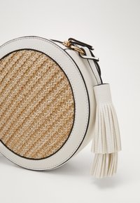Kulatá kabelka s přední stranou z pleteného přírodního vlákna a bílými koženými akcenty. Obsahuje černý okraj a detaily z bílé kožené třásně.
