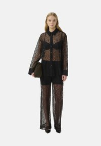 Completo in pizzo nero con una camicia trasparente con bottoni, un reggiseno aderente sotto e pantaloni coordinati. La modella tiene una borsa verde scuro.