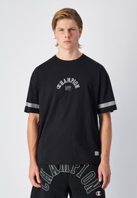Camiseta de algodón negra con cuello redondo, corte holgado y acentos de rayas grises en las mangas. Logo de texto "Champion" en un diseño curvado en la parte delantera.