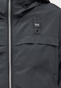 Μαύρο μπουφάν με ασημένιο φερμουάρ, κουκούλα και ορθογώνιο πατς στο στήθος που γράφει "Blauer U.S.A." με δύο μαύρες μεταλλικές τρύπες.