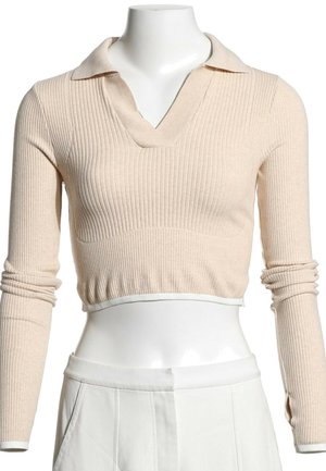 Pull court beige en maille côtelée avec manches longues et col, porté avec un pantalon blanc taille haute sur un mannequin.