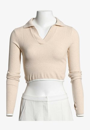 Pull court beige en maille côtelée avec manches longues et col, porté avec un pantalon blanc taille haute sur un mannequin.