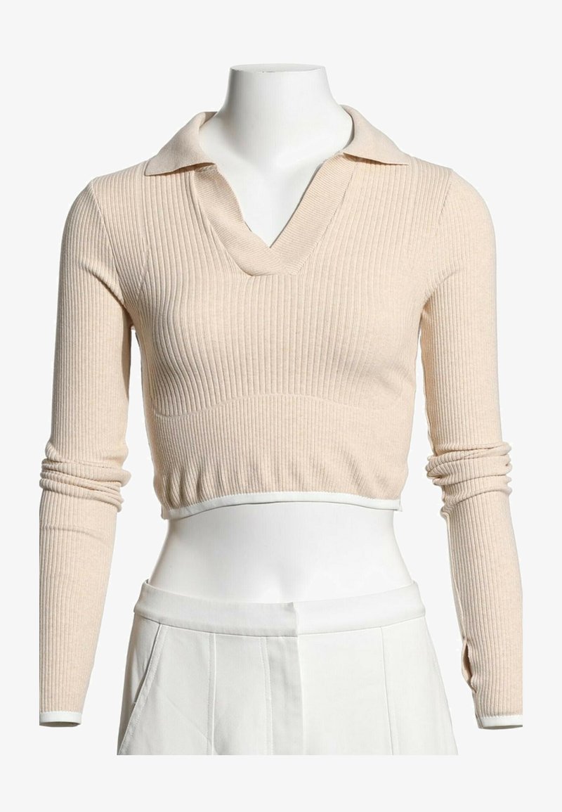 Pull court beige en maille côtelée avec manches longues et col, porté avec un pantalon blanc taille haute sur un mannequin.