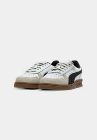 Paire de baskets basses blanches avec des bouts en suède gris, des bandes latérales noires et des semelles en caoutchouc marron sur fond blanc.
